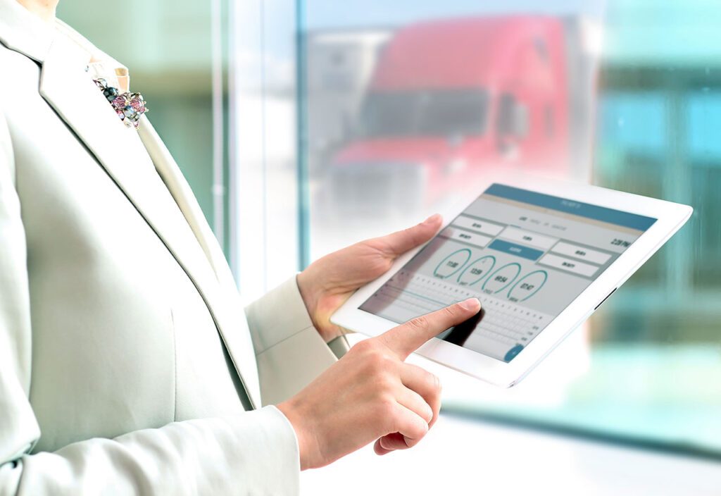 Female employee using tablet to manage trucking logistics with semi-truck in background.