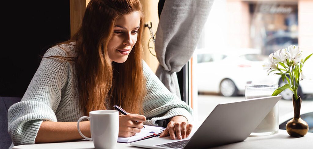 attractive woman writing in notepad while watching webinar on laptop in café