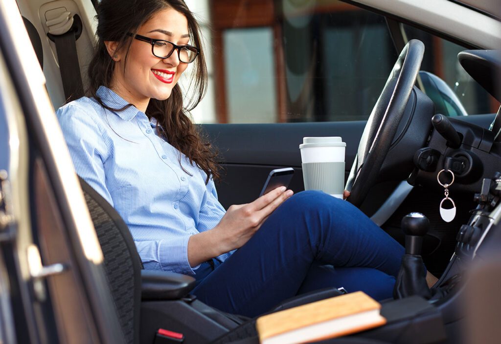 Business woman sitting in her car drinking coffee and looking at phone.