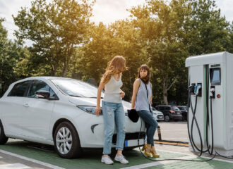 Two happy young beautiful women are talking to each other until their electric car is charging at the charging station situated in the car park.