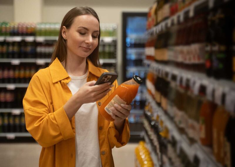 Health-conscious woman shopping in a convenience store, focusing on a peach juice among various beverages, scanning items with her phone.
