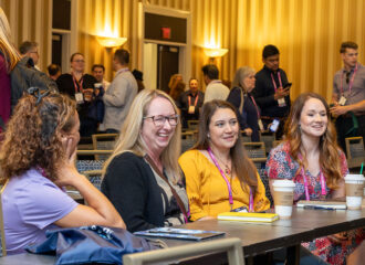 Attendees at Connections Live smiling and networking at a conference table, with notebooks and coffee cups in front of them and other participants in the background.