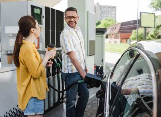 Smiling man looking at wife with coffee to go while refueling car on gas station