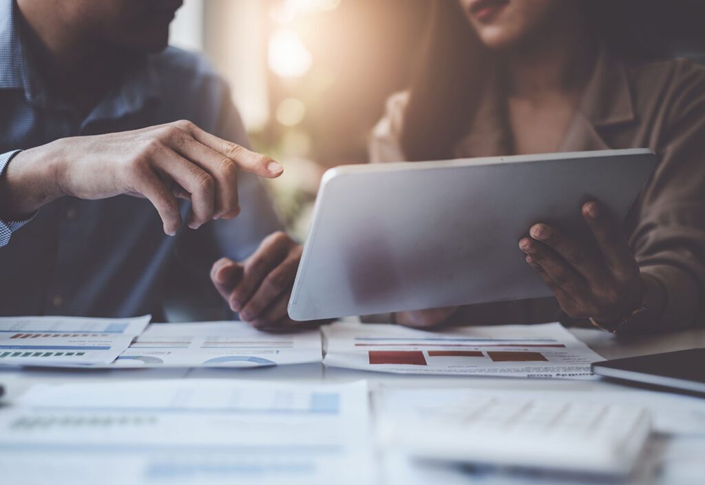Two coworkers reviewing financial data on laptop with printouts of data on desk.