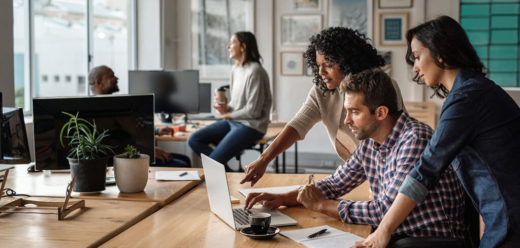 Two women and one man working together on laptop in open office environment.