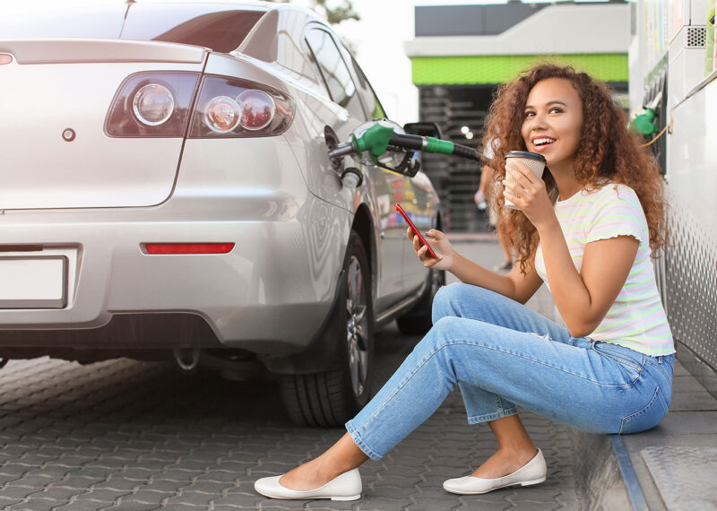 Woman sitting at rear of car near gas pumps with pump inserted into fuel tank.