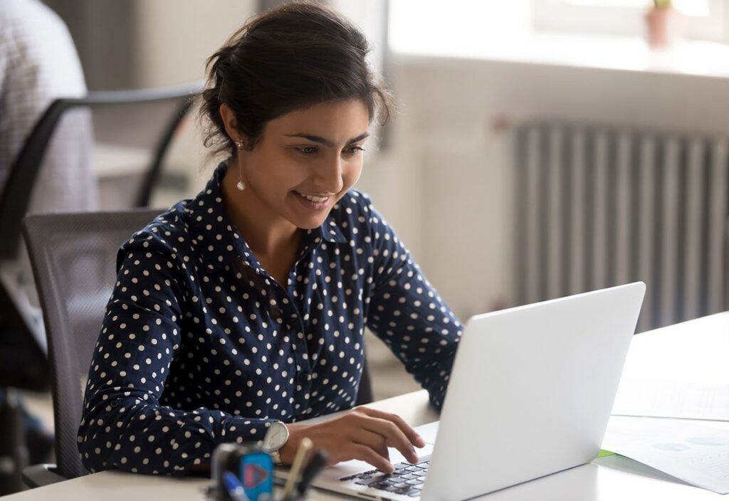 Smiling Indian female employee using laptop at workplace