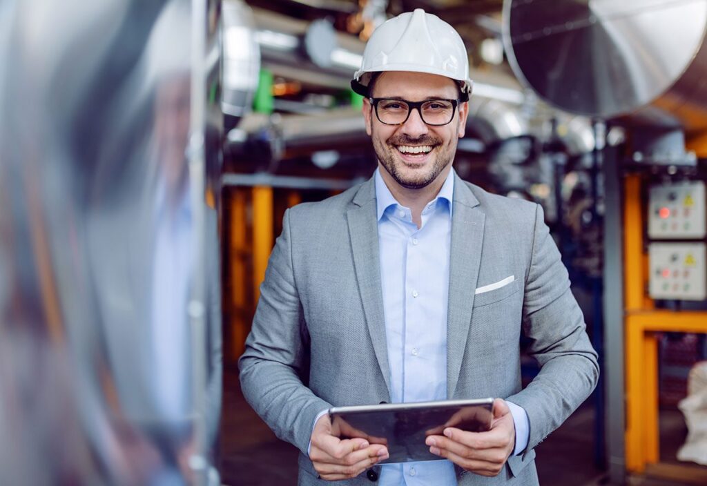 Smiling attractive Caucasian supervisor in gray suit and with white helmet on head holding tablet while standing in power plant.