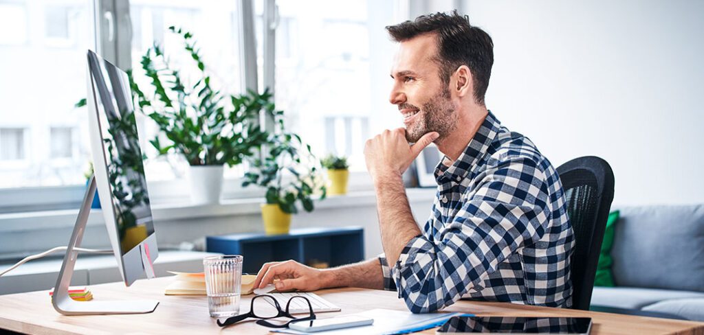 Man sitting at desk in home office smiling and working on desktop computer.