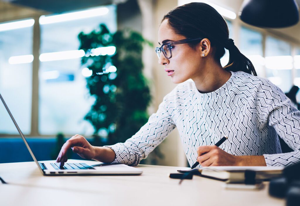 Focused woman wearing glasses using laptop while writing down information in notebook.
