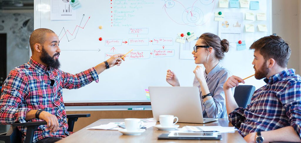 Three young coworkers reviewing business data on whiteboard using sticky notes.