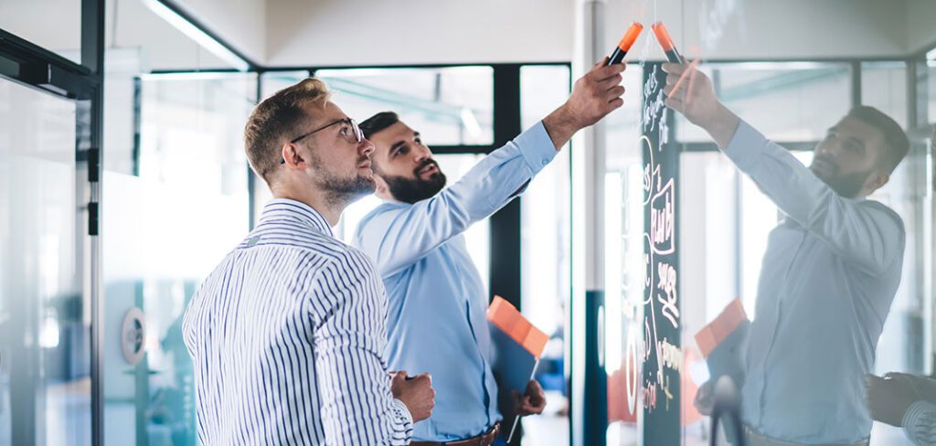 Two male businessmen writing business plans on glass wall in corporate office.