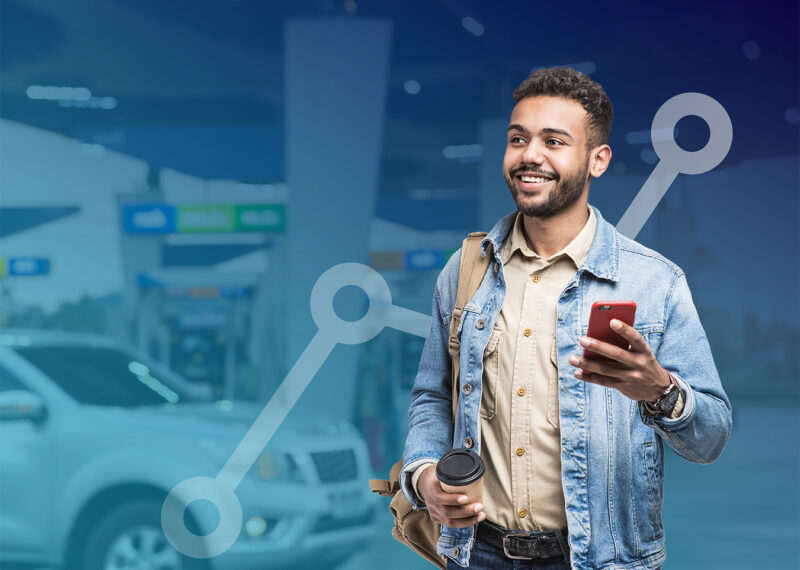 Smiling man holding coffee and mobile phone standing in front of background of convenience store forecourt with gasoline pumps