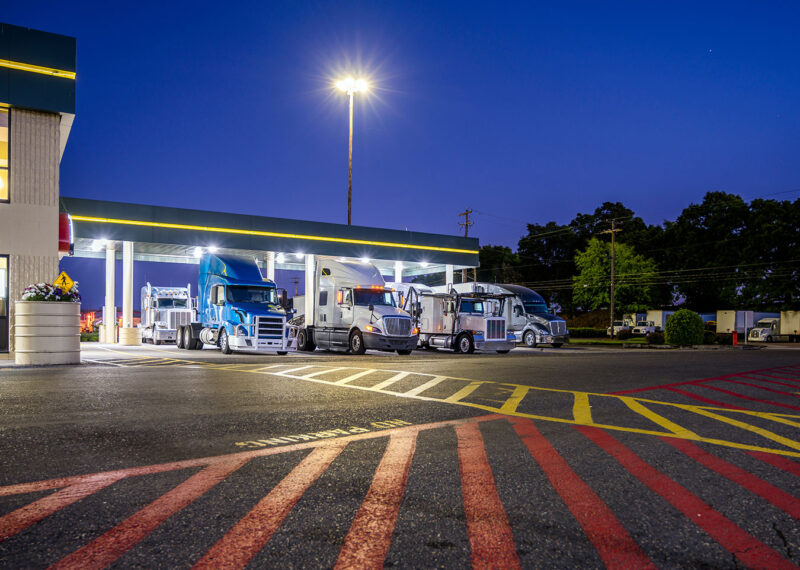 Big rigs semi trucks standing on the truck stop parking lot under a lighted awning at night