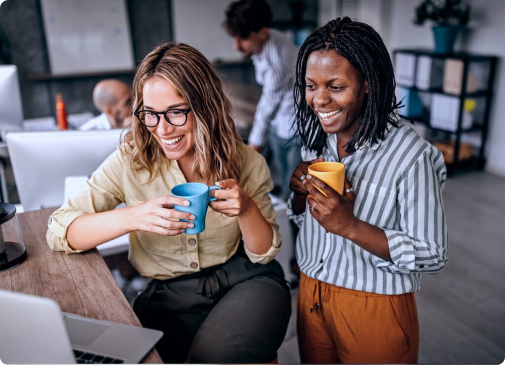 Hispanic and African American woman holding coffee cups looking at laptop in office.